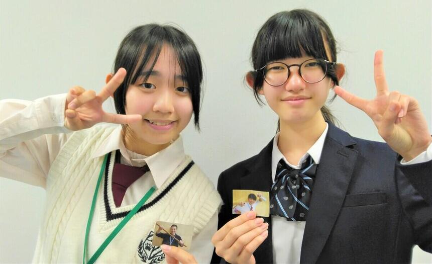 女子生徒２名が室内で男性コーチの写真を片手に持っている。こちらを見て笑顔でピースをしている。