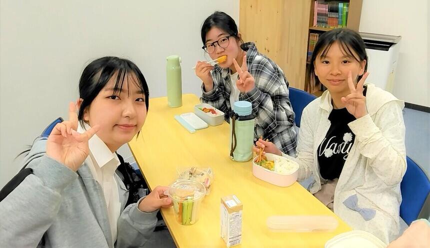 教室で3人の女子生徒がカメラ目線で食事をしながらポーズを決めている。テーブルにはお弁当箱が置かれている。