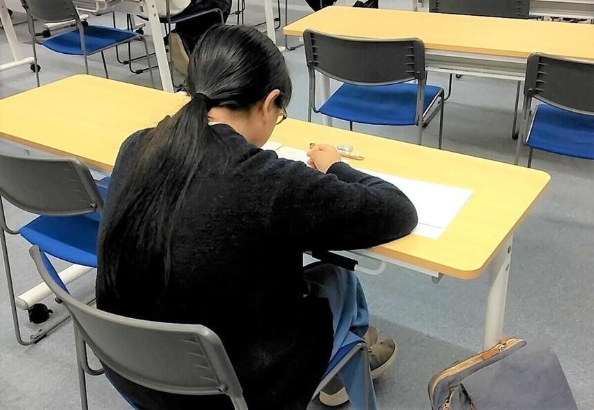一人の女子生徒がカメラに背を向けて机に向かっている。机にはプリントや筆記用具が置かれている。