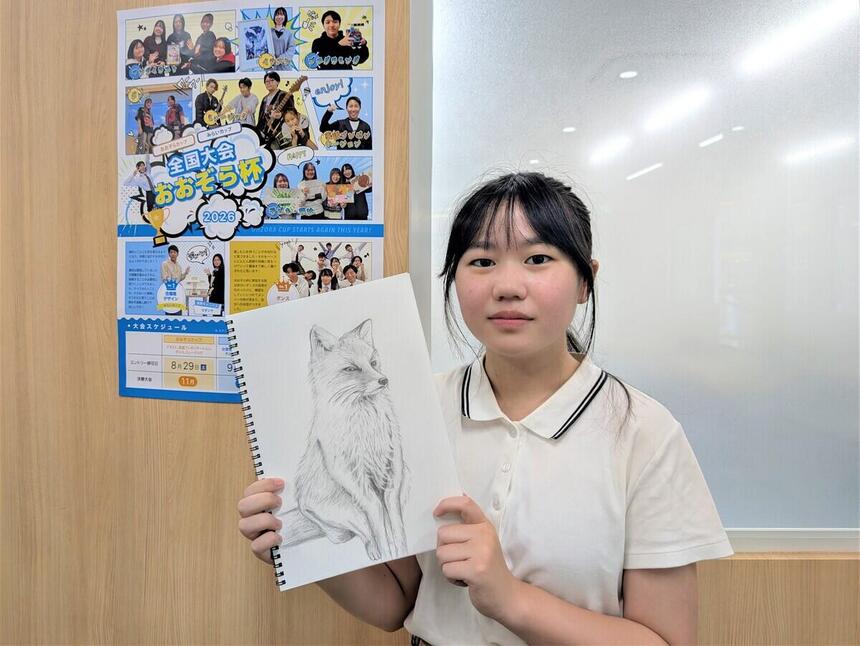 曇りガラスの窓がある壁を背景に、スケッチブックを持ってこちらを向いている女子生徒。背景には「おおぞら杯」と書かれたポスター。スケッチブックにはリアルな狐の絵が描かれている。