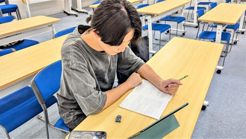 教室で一人の男子生徒が机に向かってノートに書き記している。机には消しゴム、タブレット端末、スマートフォンなどが置いてある。