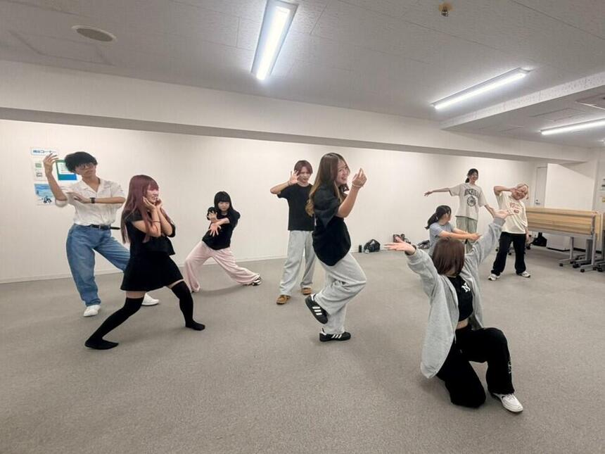 9人の男女が教室でポーズをとってこちらを向いている