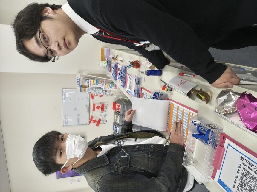 登校時のインタビュー対象生徒とマイコーチ®が登校簿を記録しながらカメラ側を見ている画像