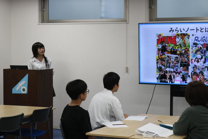 中学生に対して、在校生が教室でスライドを使って自分のみらいノートを紹介している様子