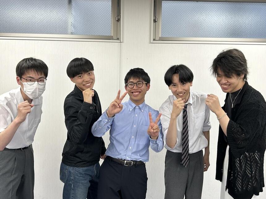 教室で男子生徒5人が制服を着てグーまたはピースのポーズをとっている写真