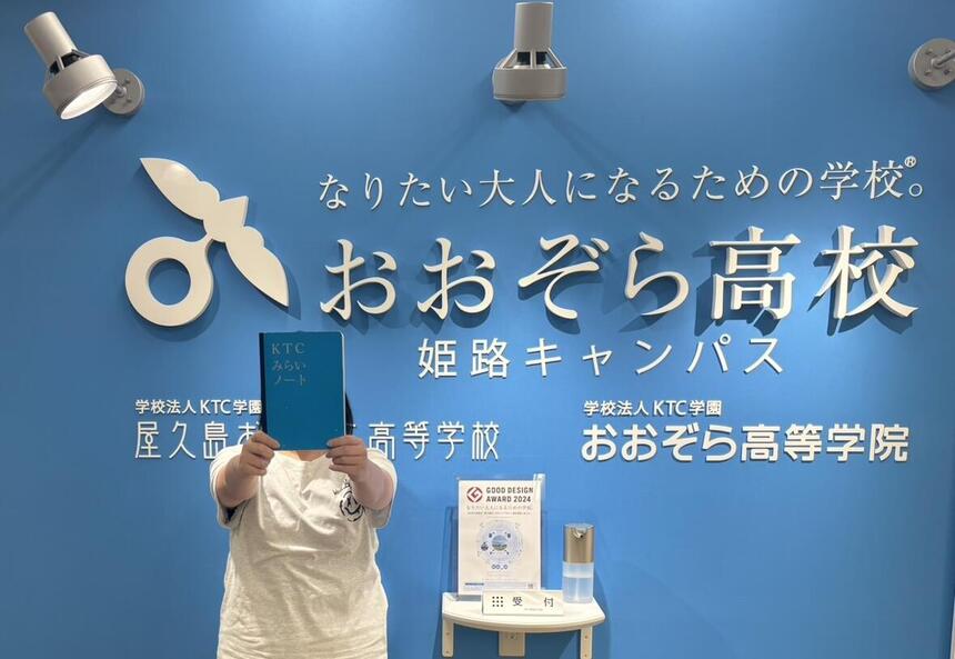 「おおぞら高校姫路キャンパス」の看板の前で、みらいノートを掲げポーズをとる姫路キャンパスの女子生徒