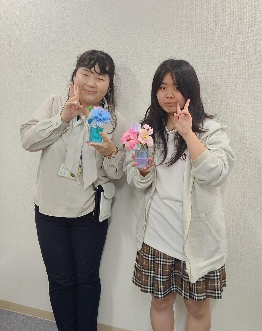 雑誌に作品が載った生徒とマイコーチが、お花（造花）をもってツーショットを撮っている写真