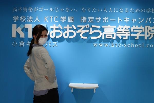 ＫＴＣおおぞら高等学院小倉キャンパス入り口の看板前で腰に手をあてて、振り向くポーズをする上之原コーチ