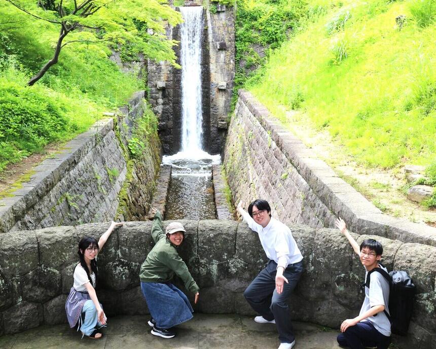 公園内の緑に囲まれて滝の前で生徒が笑顔の写真