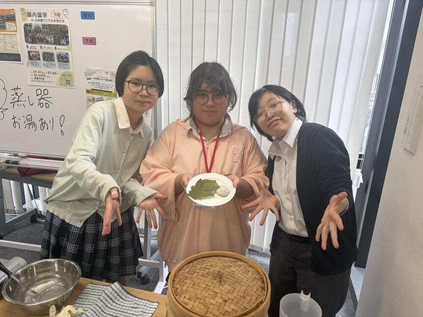 久留米キャンパスの教室で、生徒3人が「阿（ほとり）の部屋」のレッスン中に柏餅づくりを行い、完成した柏餅を笑顔で披露している様子。