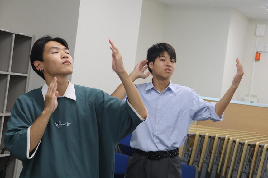 キャンパスの教室内で、2名の男子生徒が真剣な表情で両手を上げてダンスの振り付けを練習している様子