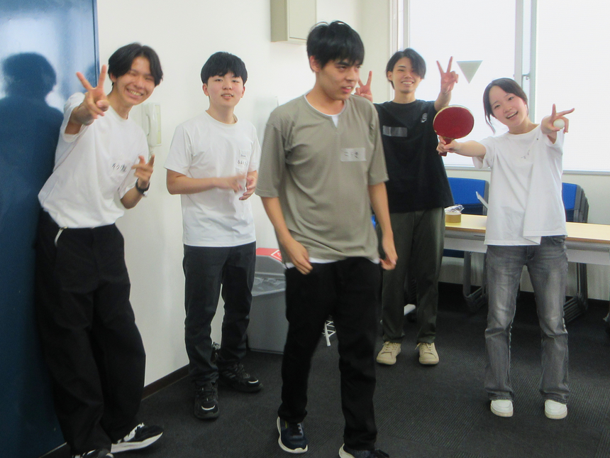 笑顔の5人の生徒が、ピースサインなどでこちらを向いている