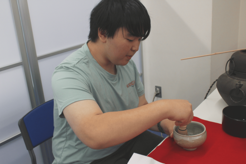 男子生徒が、机の上で茶筅を使って抹茶を点てている。室内には茶道具が並べられ、茶道体験やお茶会の準備をしている様子がうかがえる。