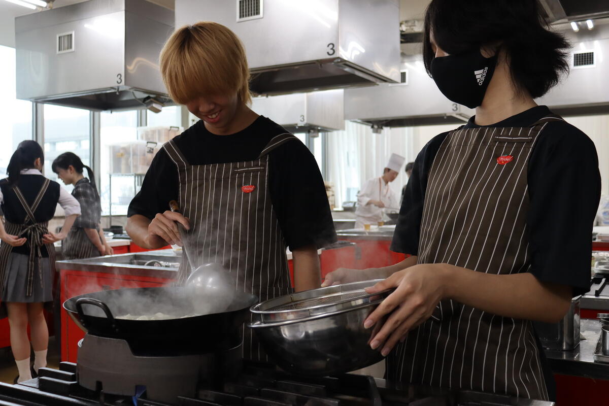  大きな網杓子（あみじゃくし）を使いこなし、火傷に気をつけながら真剣な表情で茹で加減をチェックしている生徒