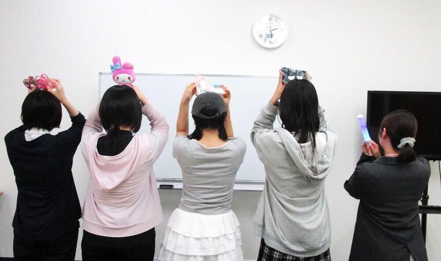 生徒とコーチが推しキャラのグッズを持って後ろ向きでポーズをとっている写真