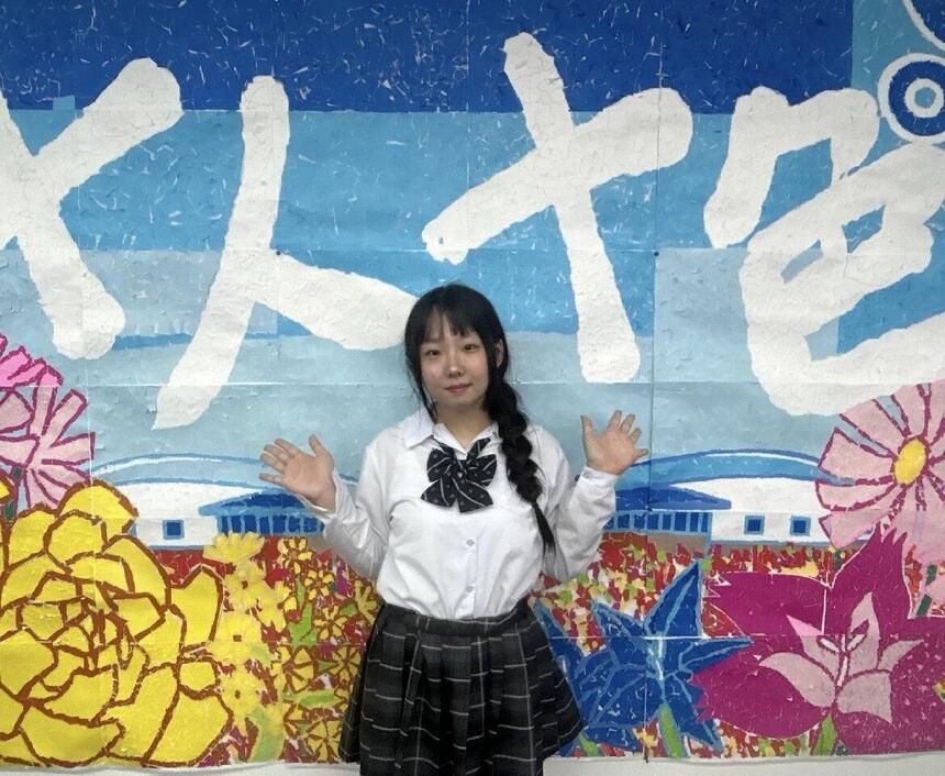 教室の壁に貼っているちぎり絵の前で、在校生のOさんが手を広げて笑顔で写っている様子。