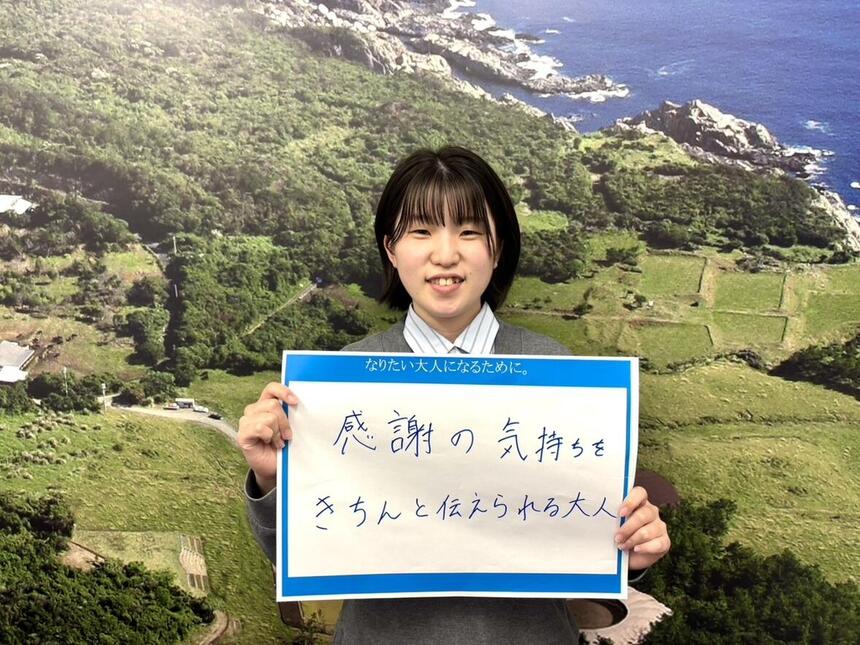 屋久島を背景にコーチの「なりたい大人」を書いた紙を持って映る女性コーチ