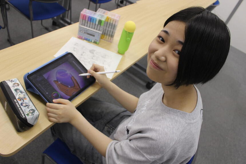 仙台キャンパスの教室で、1年次生のSさんが完成したイラスト作品を手にして笑顔で見せている様子