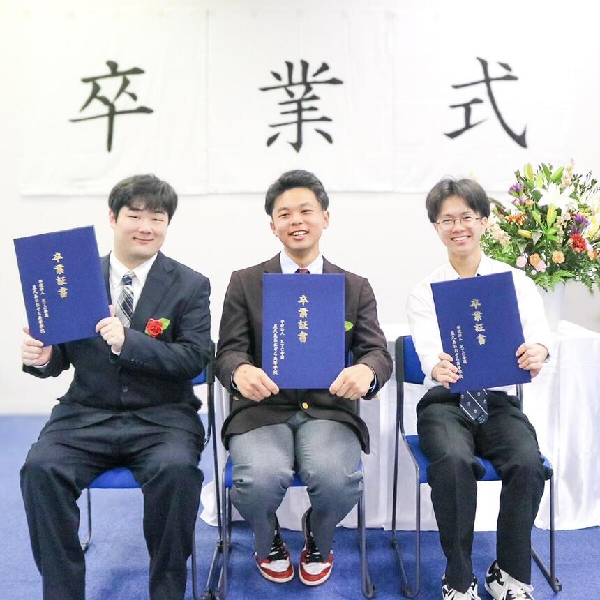 卒業式の檀上で卒業生3名が笑顔で卒業証書をもって座っている。