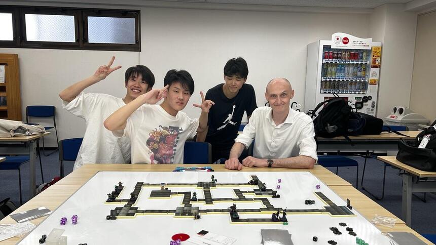 教室で、ミニチュアゲームを前に生徒と講師が集まっている様子