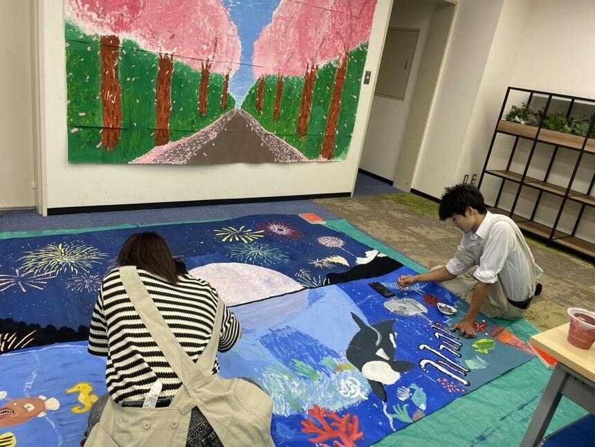 美術部員二人が大きい絵を座りながら真剣に描いている