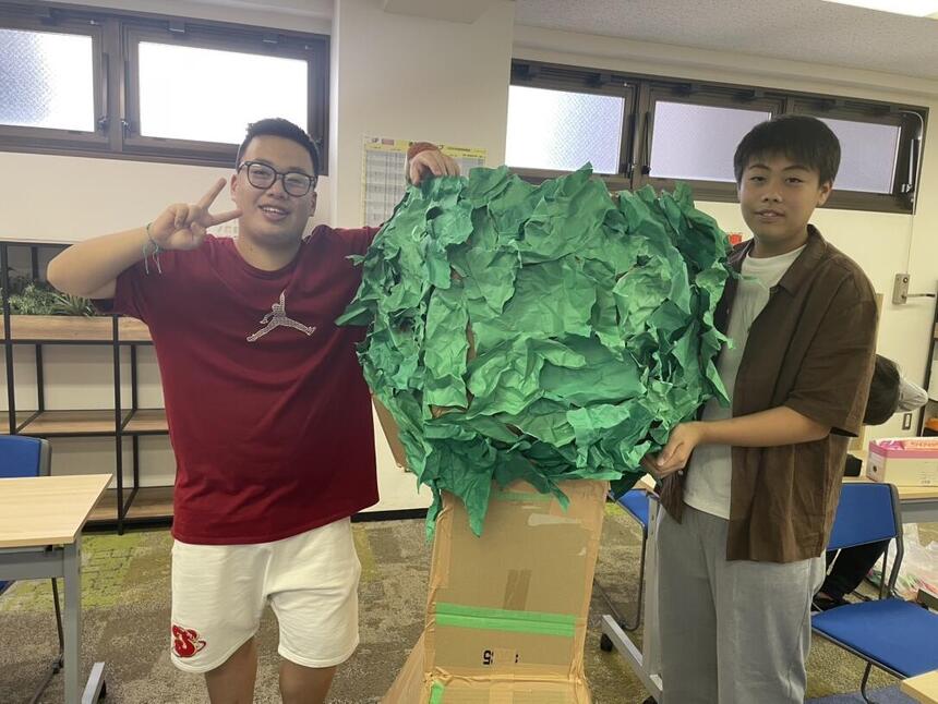 男子生徒がダンボールと紙で作った木のオブジェを笑顔で見せている画像