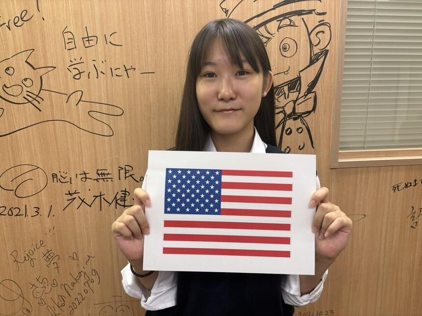 壁の前でアメリカ国旗の紙を持って、微笑えむ女子生徒