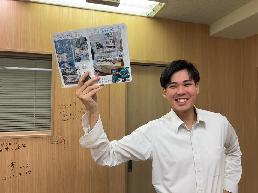 一人の男性が笑顔で、右手にノートを持って微笑んでいる写真