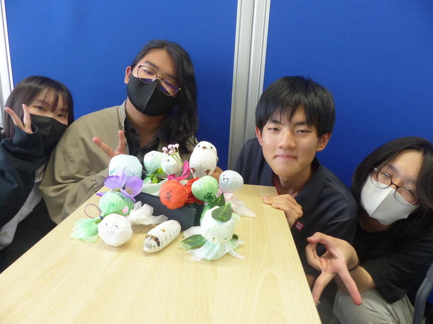 制作したてるてる坊主を机に並べ、その後ろでピースする生徒4名