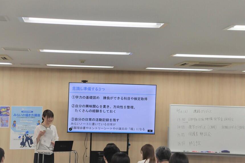 おおぞら高校の高崎キャンパスにて、進路担当の女性コーチが、大型モニターに映した「意識し準備する3つ」のスライドとホワイトボードを使い、生徒と保護者へ進路ガイダンスを行っている様子
