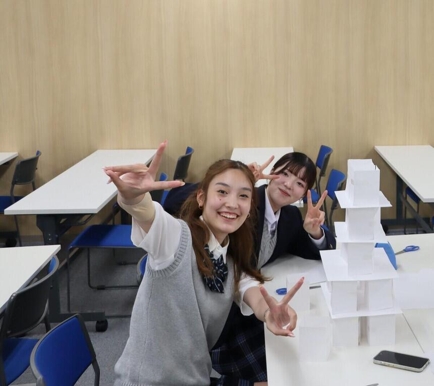 みらいの架け橋レッスン®の体験会で、高崎キャンパスの2人の女子生徒が自分たちで制作した白いペーパータワーの横で笑顔でポーズをとっている写真