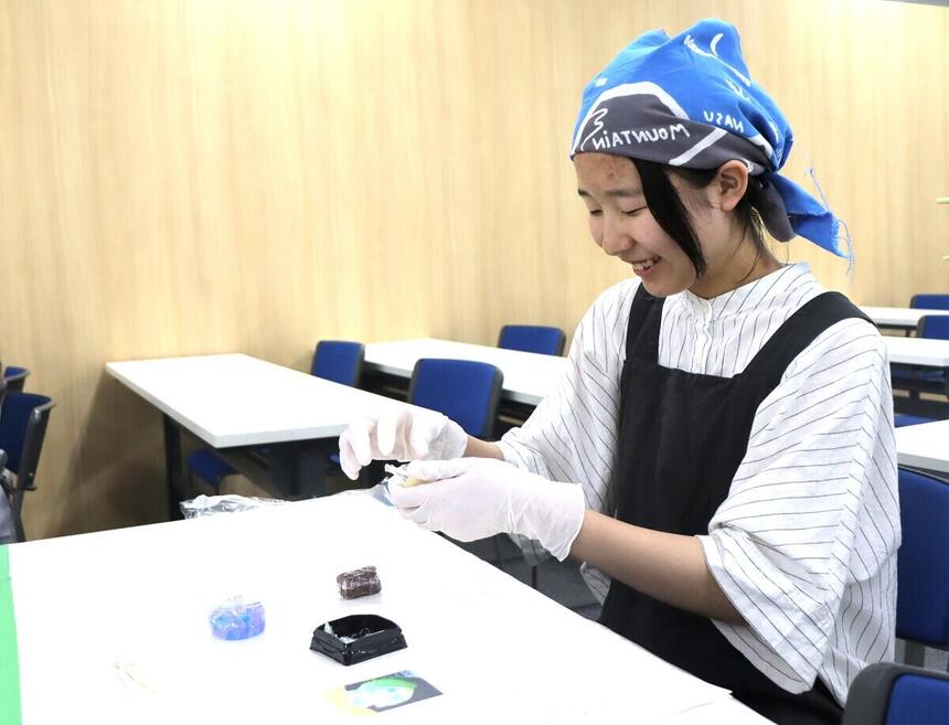 みらいの架け橋レッスン®の和菓子作り体験で、青いバンダナと黒いエプロン、白い手袋を着用した女子生徒が、机の上で笑顔でねりきりに挑戦している写真