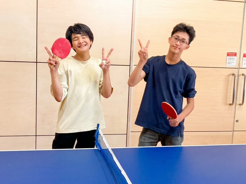 高崎アリーナの卓球台を挟んで、ラケットを持って笑顔でピースサインをするおおぞら高校高崎キャンパスの男子生徒2人