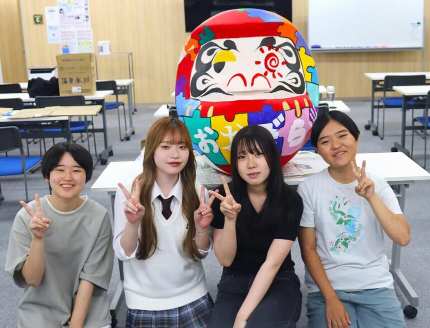 高崎キャンパスの中で完成したカラフルなパズル柄のだるまの前で、ピースサインで笑顔を見せる高崎キャンパスの生徒4人