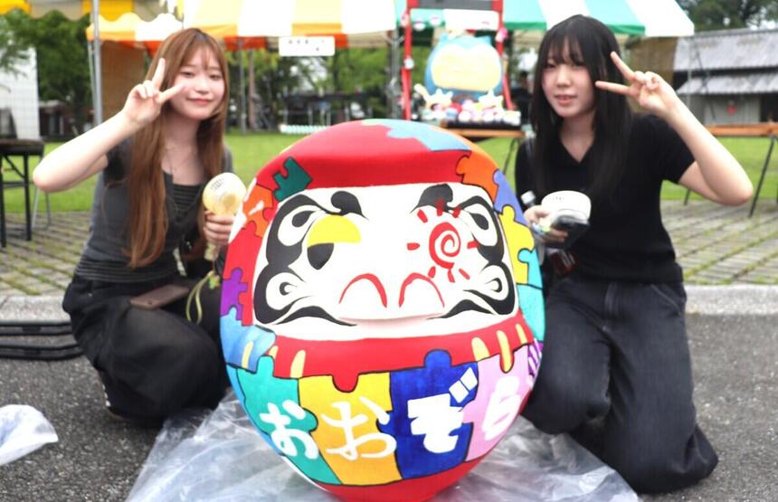 33℃の猛暑の中、完成した特大だるまの前で手持ち扇風機を持ち笑顔でダブルピースをする2人の女子生徒