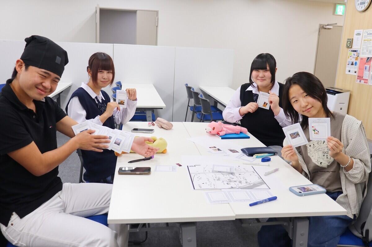 高崎キャンパスの机を囲み、完成したおすすめスポットカードを笑顔で見せ合う4人の生徒