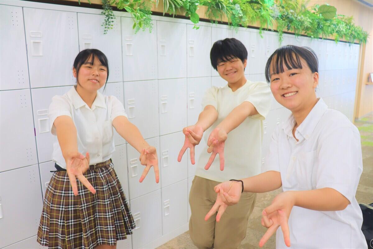 白いロッカーの前で夏休み明けに再会した3人の生徒が笑顔で「高崎だるま」の手ポーズをしてポーズをとっている様子