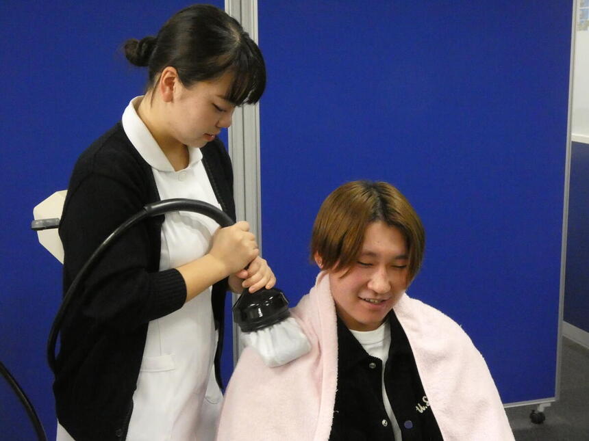 専門学校の生徒が振動する機械を生徒にあてています。生徒は気持ちよさそうです。