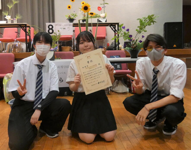 宇都宮キャンパスの3人の生徒たちが、花の甲子園®で作り上げた生け花作品の前で賞状を手にしています。