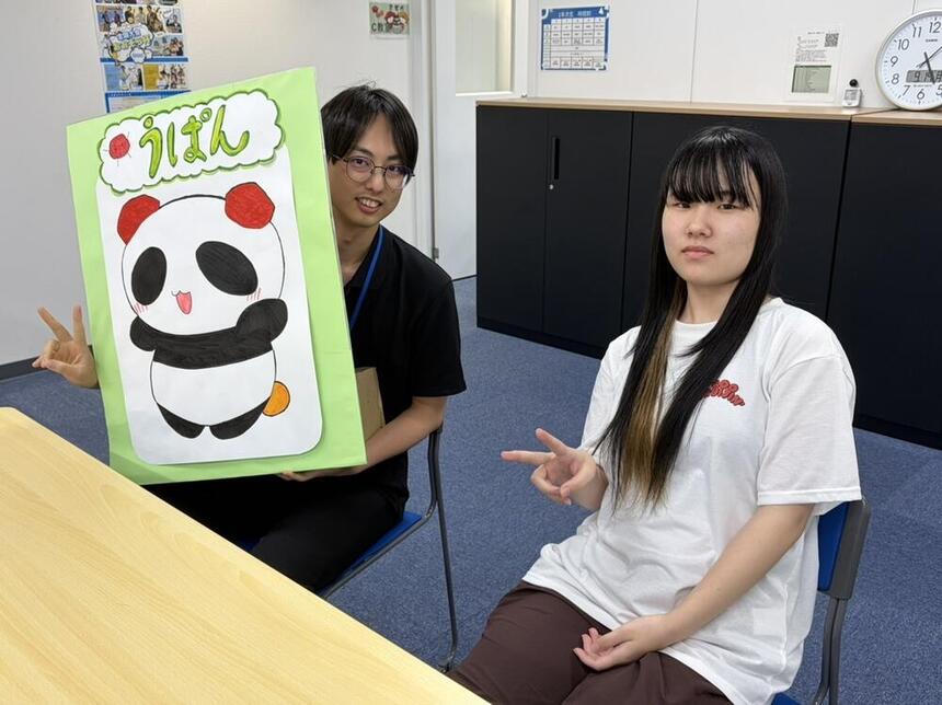 和歌山キャンパスオリジナルキャラクターの看板を持つマイコーチ®とすてきな笑顔で写真に写るSさん