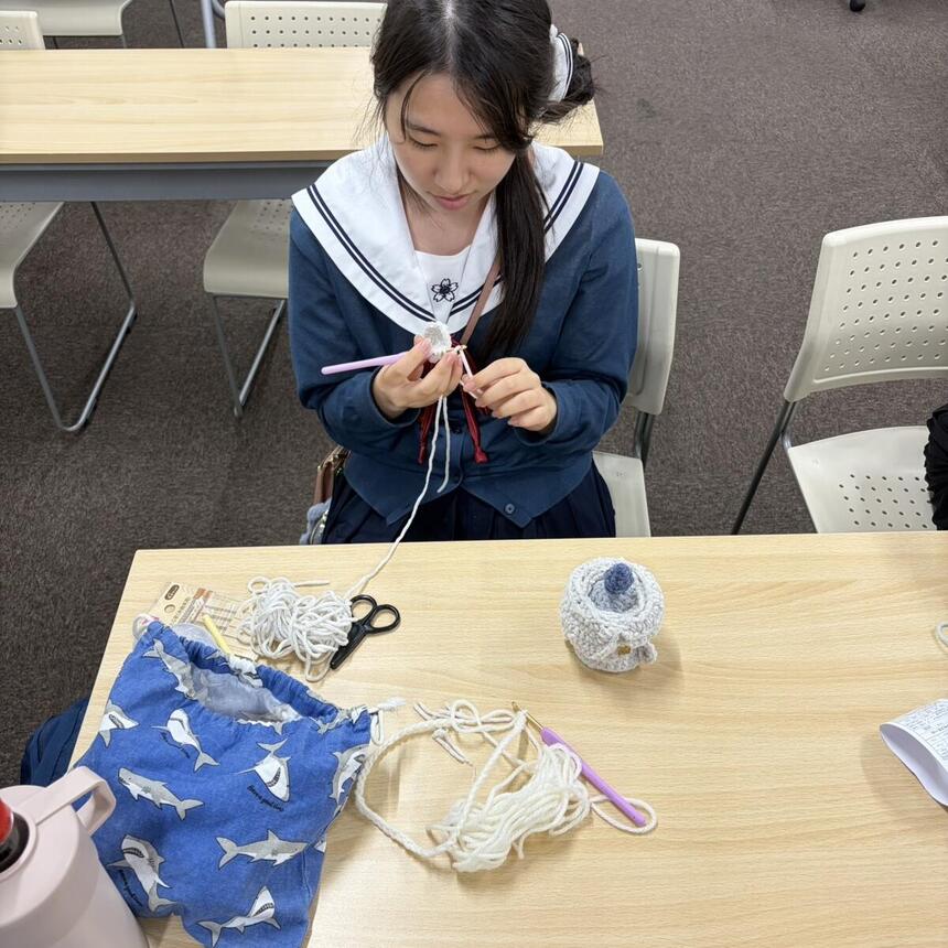 生徒が編み物をしている様子。机の上には毛糸の束と、コーチが作成した鎖編みの作品がある。