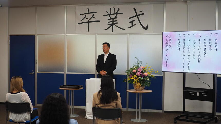 卒業式の釣り看板の下で卒業生に向けて話すキャンパス長