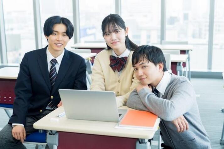 学生服をした男子生徒2人と女子生徒1人がパソコンが置かれた机を囲みながら微笑んでいる