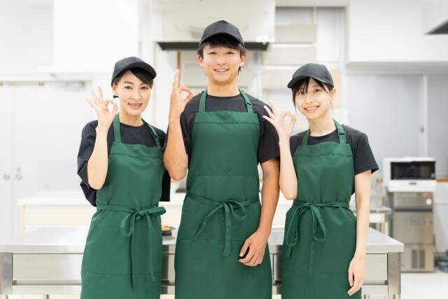 調理場で緑のエプロンをつけたアルバイトスタッフ3人がポーズを笑顔でOKポーズをとっている