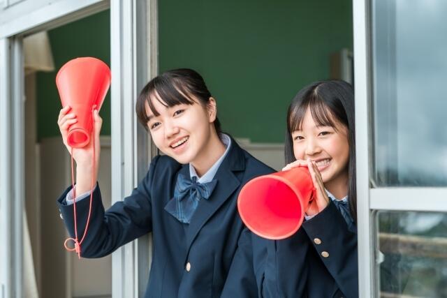 制服を着ている女子生徒2人が赤いメガホンを持って校舎の窓から応援している