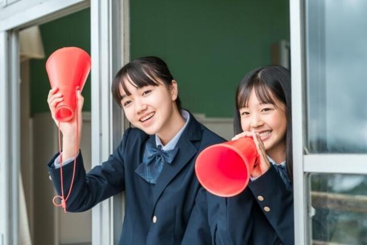 制服を着ている女子生徒2人が赤いメガホンを持って校舎の窓から応援している