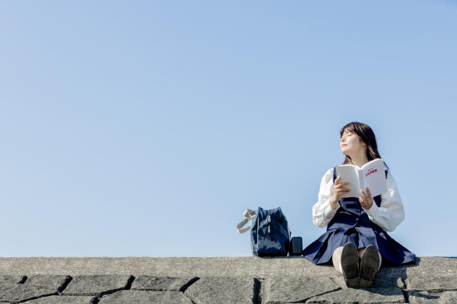 土手で制服を着た女子生徒が本を読みながら空を見上げている様子