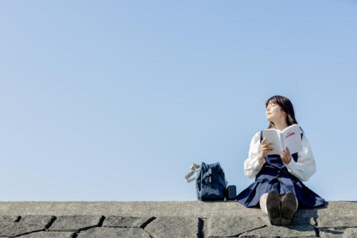 土手で制服を着た女子生徒が本を読みながら空を見上げている様子