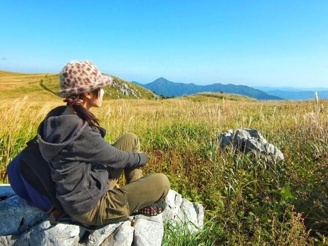 帽子をかぶった女性が草原で石の上に座って景色を眺めている様子