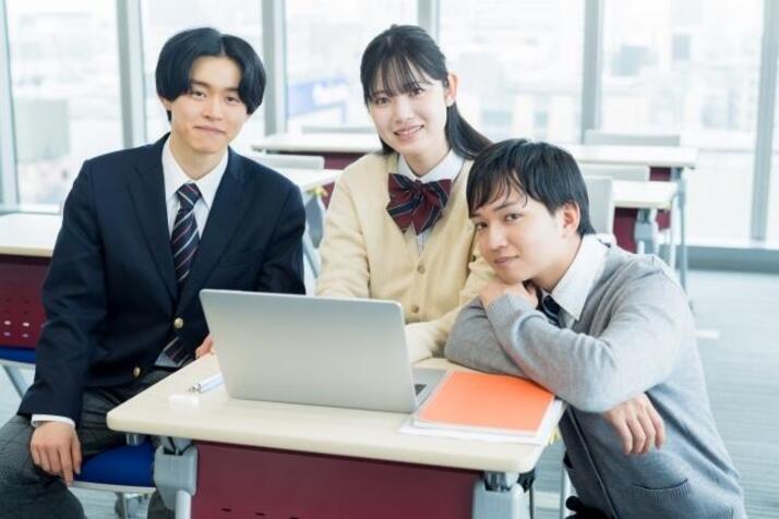 3人の生徒たちがノートパソコンが置いてある勉強机の前でカメラ目線で写真に写っている様子